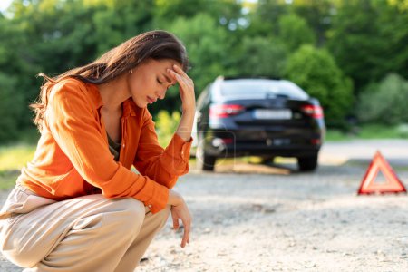Traurige junge Fahrerin sitzt auf Straße mit kaputtem Auto im Hintergrund, aufgebrachte Dame wartet auf Hilfe, hat Probleme mit Fahrzeug