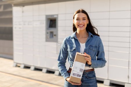 Happy lady holding mailbox in her hands, standing near automatic post terminal and smiling at camera, free space. Concept of smart delivery, modern shipping and logistics