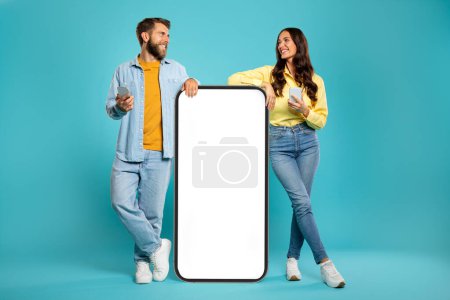Stylish man and woman using phones and looking at each other, standing near huge smartphone with white empty screen on blue studio background, mockup