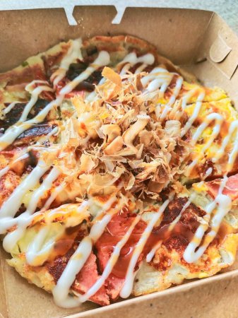Typischer japanischer Straßensnack, Okonomiyaki in einer Verpackungsbox. Okonomiyaki wird aus Weizenmehl hergestellt, das Gemüse, Fleisch, Fisch usw. enthält. Dann auf einer Eisenplatte gebacken und mit Sauce beschmiert