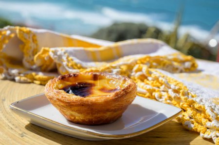 Photo for Portugal's traditional sweet dessert Pastel de nata egg custard tart pastry served with view on blue Atlantic ocean in Lisbon area, Portugal, close up - Royalty Free Image