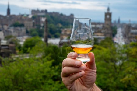 Foto de Hand holding glass of single malt scotch whisky and view from Calton hill to park and old parts of Edinburgh city in rainy summer day, Scotland, UK - Imagen libre de derechos