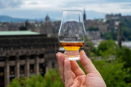 Hand holding glass of single malt scotch whisky and view from Calton hill to park and old parts of Edinburgh city in rainy summer day, Scotland, UK