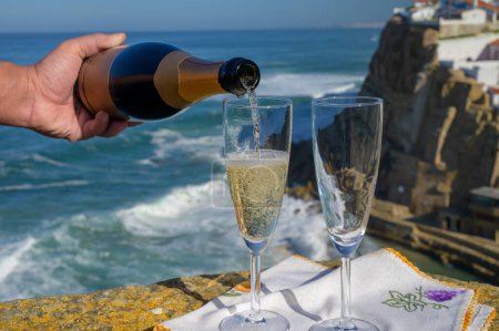 Téléchargez les photos : Verser du champagne mousseux vin blanc dans des verres avec vue sur les maisons blanches du village pittoresque Azenhas do mar, Lisbonne, Portugal - en image libre de droit