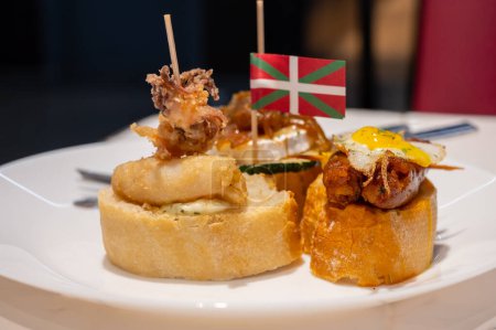 Weißes Brett mit typischen Snacks und Flagge des Baskenlandes, Pinchos oder Pinxtos Spieße mit kleinen Brotstücken, Zucchini, Meeresfrüchten, Eiern, Käse, serviert in einer Bar in San-Sebastian oder Bilbao, Spanien