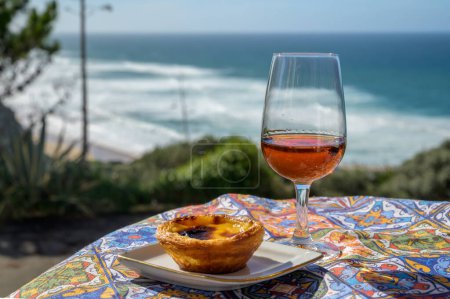 Portugals traditionelles Essen und Trinken, Glas Porto Wein oder Muscatel de setubal, süßes Dessert Pastel de nata Eierpudding Torte mit Blick auf den blauen Atlantik in der Nähe von Sintra im Raum Lissabon, Portugal