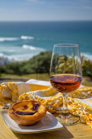 Portugal's traditional food and drink, glass of porto wine or muscatel de setubal, sweet dessert Pastel de nata egg custard tart pastry served with view on blue Atlantic ocean near Sintra in Lisbon area, Portugal