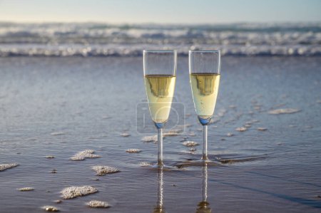 Téléchargez les photos : Lunettes de cava ou champagne mousseux sur plage de sable blanc avec des vagues d'eau au coucher du soleil dans les rayons du soleil avec des ombres - en image libre de droit