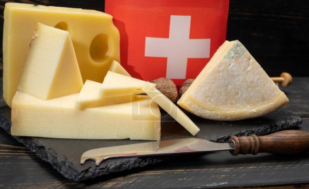 Téléchargez les photos : Assortiment de fromages suisses Emmental ou Emmentaler fromage moyen à pâte dure avec trous ronds, Gruyere, appenzeller utilisé pour la fondue et le gratin de fromage traditionnel et drapeau de Suisse sur fond sombre - en image libre de droit