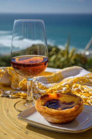 Photo for Portugal's traditional food and drink, glass of porto wine , sweet dessert Pastel de nata egg custard tart pastry served with view on blue Atlantic ocean near Sintra in Lisbon area Portugal - Royalty Free Image