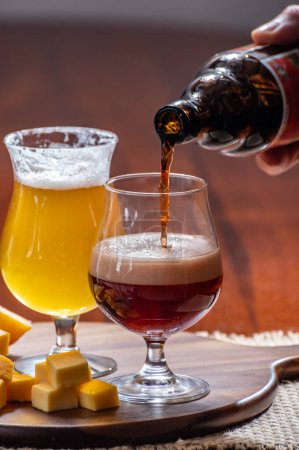 Glasses of Belgian yellow blond and dark brown beer made in abbey and wooden board with variety of belgian cheeses, food and beer pairing in Belgium