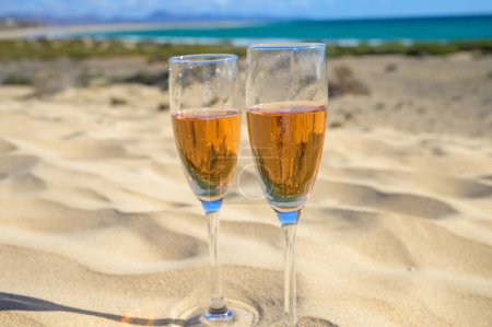 Téléchargez les photos : Two glasses of rose champagne or cava sparkling wine served on the white sandy tropical beach with dunes and blue ocean, romantic vacation, winter sun on Fuerteventura, Canary, Spain - en image libre de droit