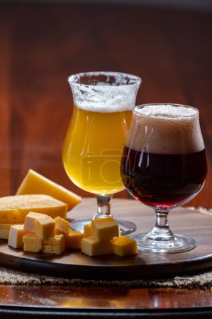 Foto de Vasos de cerveza rubia amarilla belga y marrón oscuro hechos en abadía y tablero de madera con variedad de quesos belgas, comida y maridaje de cerveza en Bélgica - Imagen libre de derechos