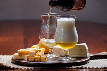 Verter cerveza rubia ligera belga hecha en abadía y tablero de madera con variedad de quesos belgas, comida y maridaje de cerveza