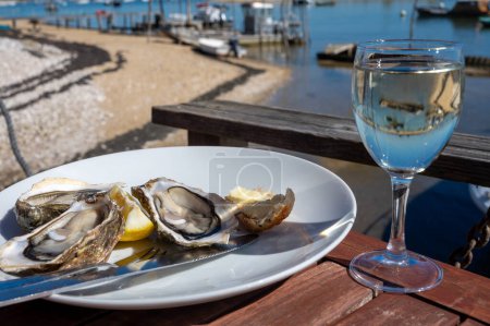 Eating of fresh live oysters with glass of white wine at farm cafe in oyster-farming village, with view on boats and water of Arcachon bay, Cap Ferret peninsula, Bordeaux, France in sunny day