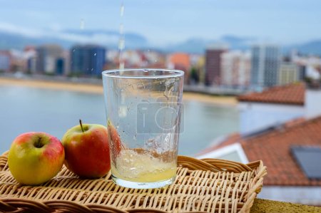 Téléchargez les photos : Cidre asturien naturel traditionnel fabriqué à partir de pommes fermentées en barrique de bois doit être versé de grande hauteur pour bulles d'air dans la boisson et vue sur la plage de San Lorenzo de Gijon - en image libre de droit