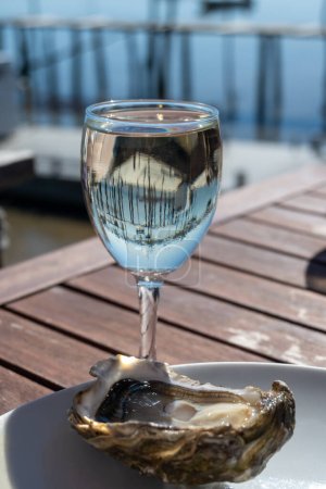 Photo for Eating of fresh live oysters with glass of white wine at farm cafe in oyster-farming village, with view on boats and water of Arcachon bay, Cap Ferret peninsula, Bordeaux, France in sunny day - Royalty Free Image