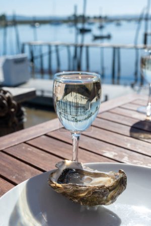 Photo for Eating of fresh live oysters with glass of white wine at farm cafe in oyster-farming village, with view on boats and water of Arcachon bay, Cap Ferret peninsula, Bordeaux, France in sunny day - Royalty Free Image
