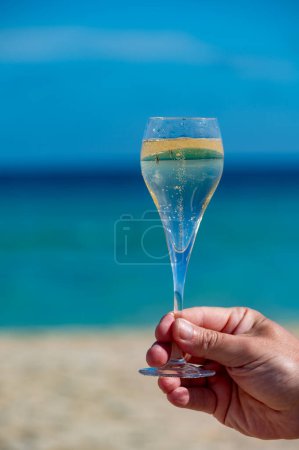 Hand with glass of cava or champagne sparkling wine on vacation, Dunes Corralejo Grand sandy beach, Fuerteventura, Canary islands, blue ocean