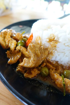 Foto de Receta de pollo al curry (Panang Kai) con arroz jazmín en plato negro sobre mesa de madera. Cocina tailandesa Curry con cacahuetes, coco y carne. Comida rápida estilo tailandés - Imagen libre de derechos