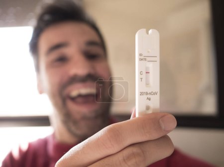 Photo for Man holds negative COVID test, out of focus, joyful with mouth open, at home by the window. A candid moment capturing relief and happiness - Royalty Free Image