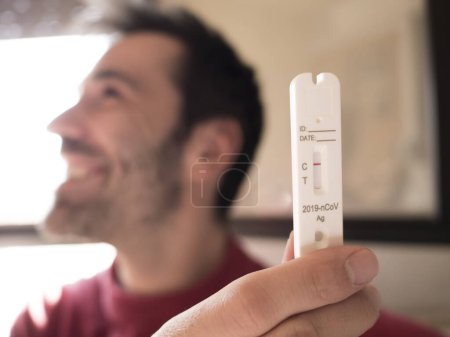 Photo for Optimistic scene: A man with black hair and beard displays a negative COVID test, exuding happiness. Soft focus captures his joyful gaze by the window - Royalty Free Image