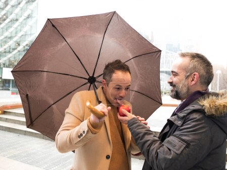 executive extends a red apple to a colleague who accepts it under an umbrella on a rainy day. The colleague begins to enjoy the apple, their shared shelter keeping them dry