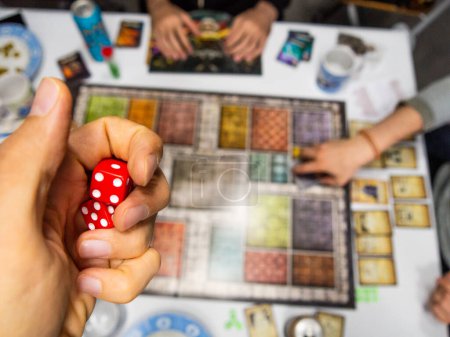 Hand with red six-sided dice on game board from above with other real players one four and five cards