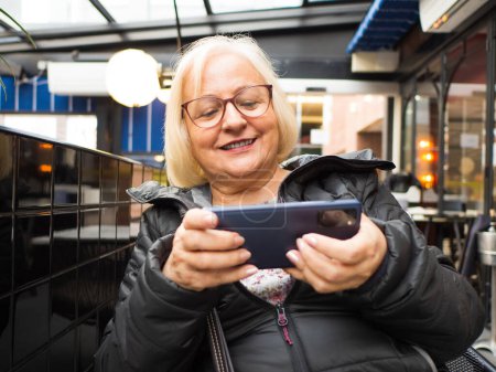 Lächelnde blonde Spielergroßmutter, die auf einer Restaurantterrasse mit dem Handy spielt und das Spiel gewinnt