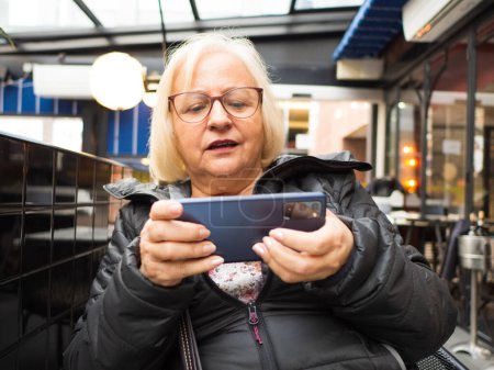 Blonde Spielergroßmutter, die auf einer Restaurantterrasse am Handy spielt, verliert das Spiel