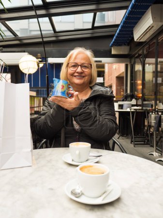 Lächelnde blonde Seniorin zeigt handgemachtes Krug-Geschenk zum Muttertag im Café