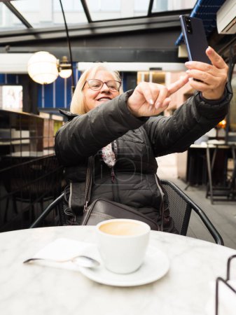 Die blonde Seniorin mit der Brille lacht und macht ein Selfie mit dem Handy, während der Kaffee schon fertig ist