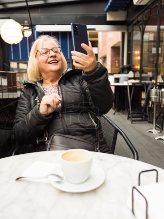 pov senior blonde woman with glasses laughing watching selfie with the cell phone with the coffees already finished