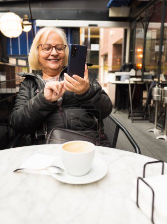 pov mujer rubia mayor con gafas riendo enviando selfie en el teléfono celular con los cafés ya terminados