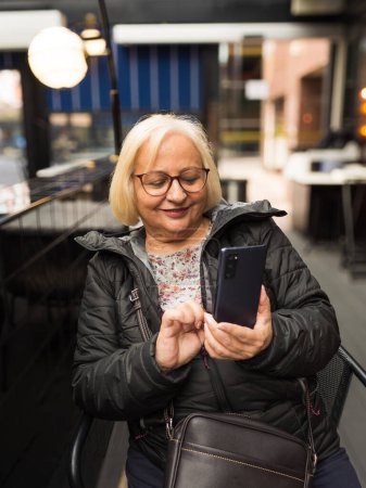 rubia mujer mayor con gafas feliz mirando el teléfono celular en un café