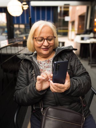 Blonde Seniorin mit Brille berührt glücklich Handy in Café