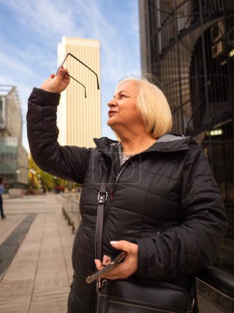 Blonde Geschäftsfrau blickt im Winter im Büroviertel mit Handy in der Hand auf ihre Brille