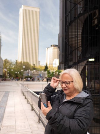 senior blonde business woman looks at cell phone holding glasses checking her vision on the street in the daytime in winter