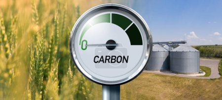 Gauge with inscription CARBON. Arrow points to zero. Agricultural field and silos on a background. Concept of carbon neutral bio fuel 