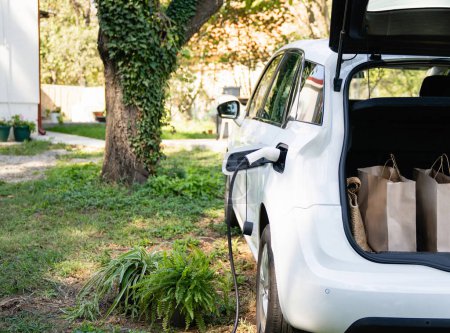 Man with shopping bag next to a charging electric car in the yard of a country house.