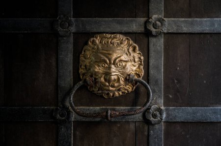 Foto de Golpeador de puerta rústico en forma de cabeza de león, pintado de oro, en una antigua puerta de madera medieval - Imagen libre de derechos