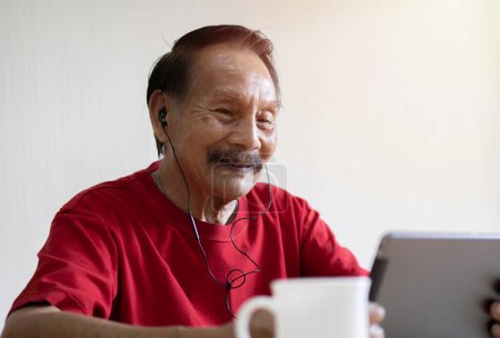 Foto de Retrato de un hombre asiático feliz usando taplet con auriculares mientras está sentado en el escritorio en el descanso del café. Concepto de estilo de vida jubilatorio. - Imagen libre de derechos