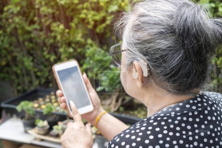 Foto de Primer plano de la anciana asiática que usa audífono en la oreja mientras usa un teléfono inteligente con fondo natural en casa. Enfoque selectivo, espacio de copia - Imagen libre de derechos