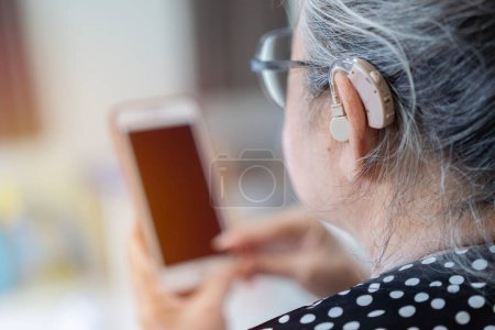 Foto de La mujer adulta con discapacidad auditiva ajusta la configuración de su audífono a través de un teléfono inteligente. Audífonos, tratamiento de sordera, tecnologías innovadoras en audiología - Imagen libre de derechos