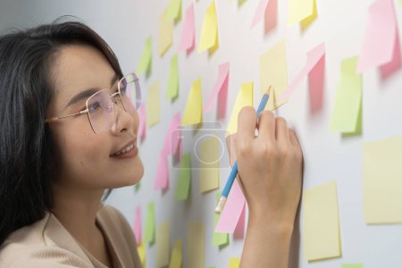 Foto de Feliz mujer de negocios asiática escribir una breve nota para hacer la lista de pegatinas en la pared en la sala de reuniones oficina. Enfoque sectorial. - Imagen libre de derechos