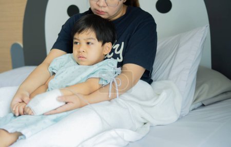 Cropped shot of Asian mother holding hand of tired little son patient lying in hospital bed with saline solution.