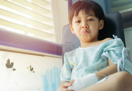 Portrait of sick little Asian boy feeling worry with saline solution sitting on chair by the window in hospital bed