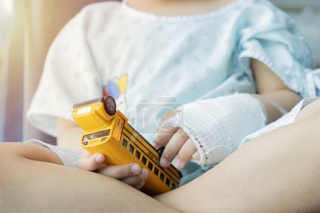 Sick kid in hospital with intravenous (IV) a saline on hand and holding a toy car while sitting in a chair by the window. Selective focus.