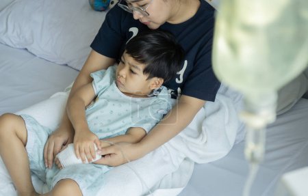 The mother is supporting her son while lying sick and giving medicine through the brine line in the patient's bed with concern in the hospital.