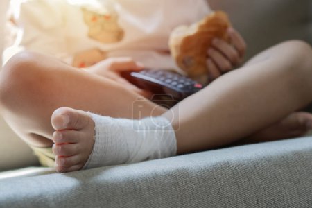 Kids healthcare and medicine concept - little child boy siting on sofa with plaster bandage on leg. Selective focus.